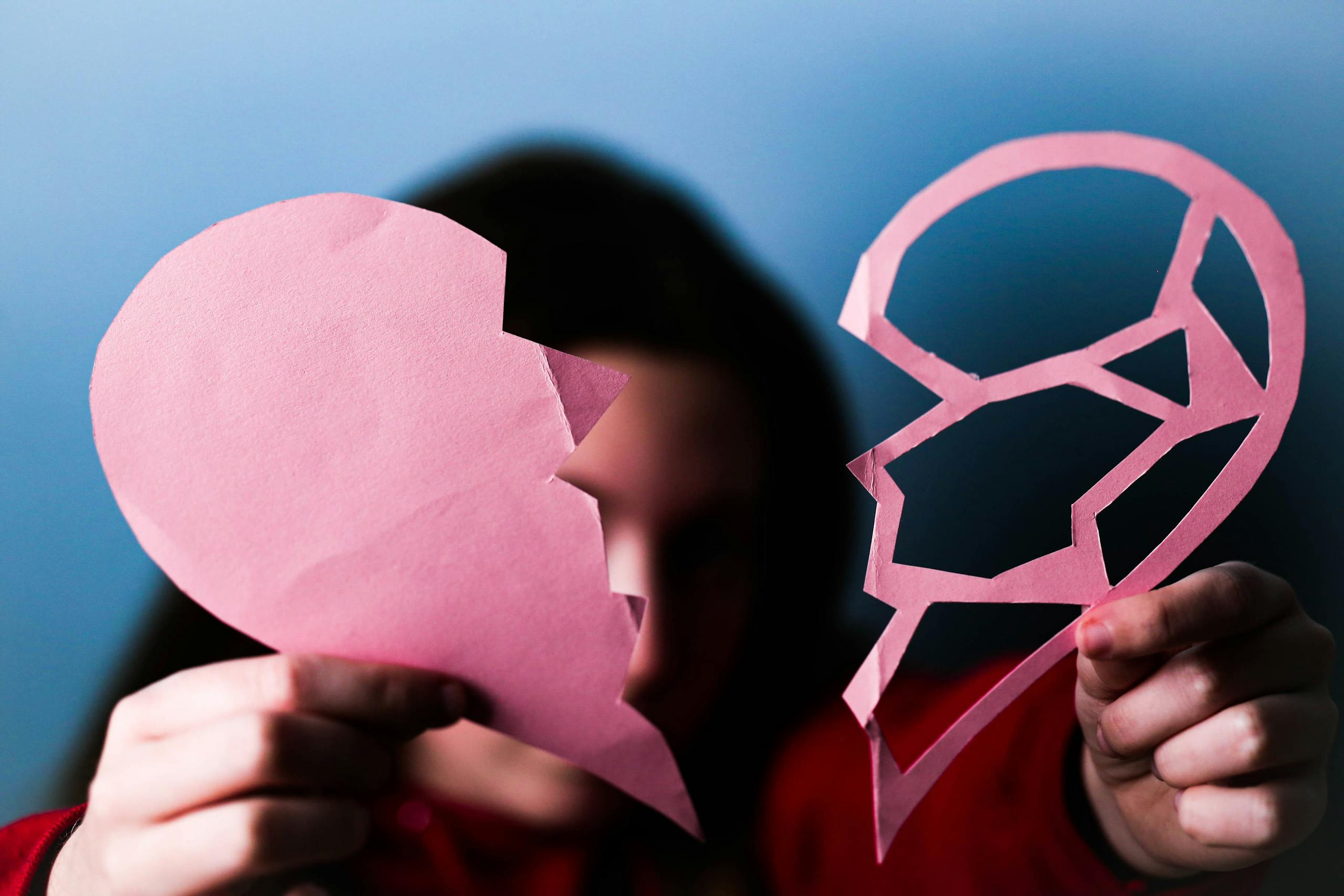 Close-up of hands holding a pink broken paper heart, symbolizing love and heartbreak.